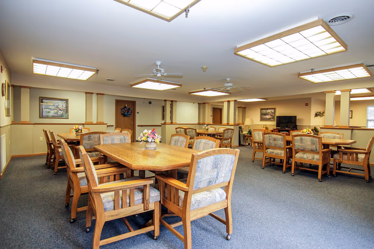 A spacious senior living dining hall with multiple wooden tables and upholstered chairs.