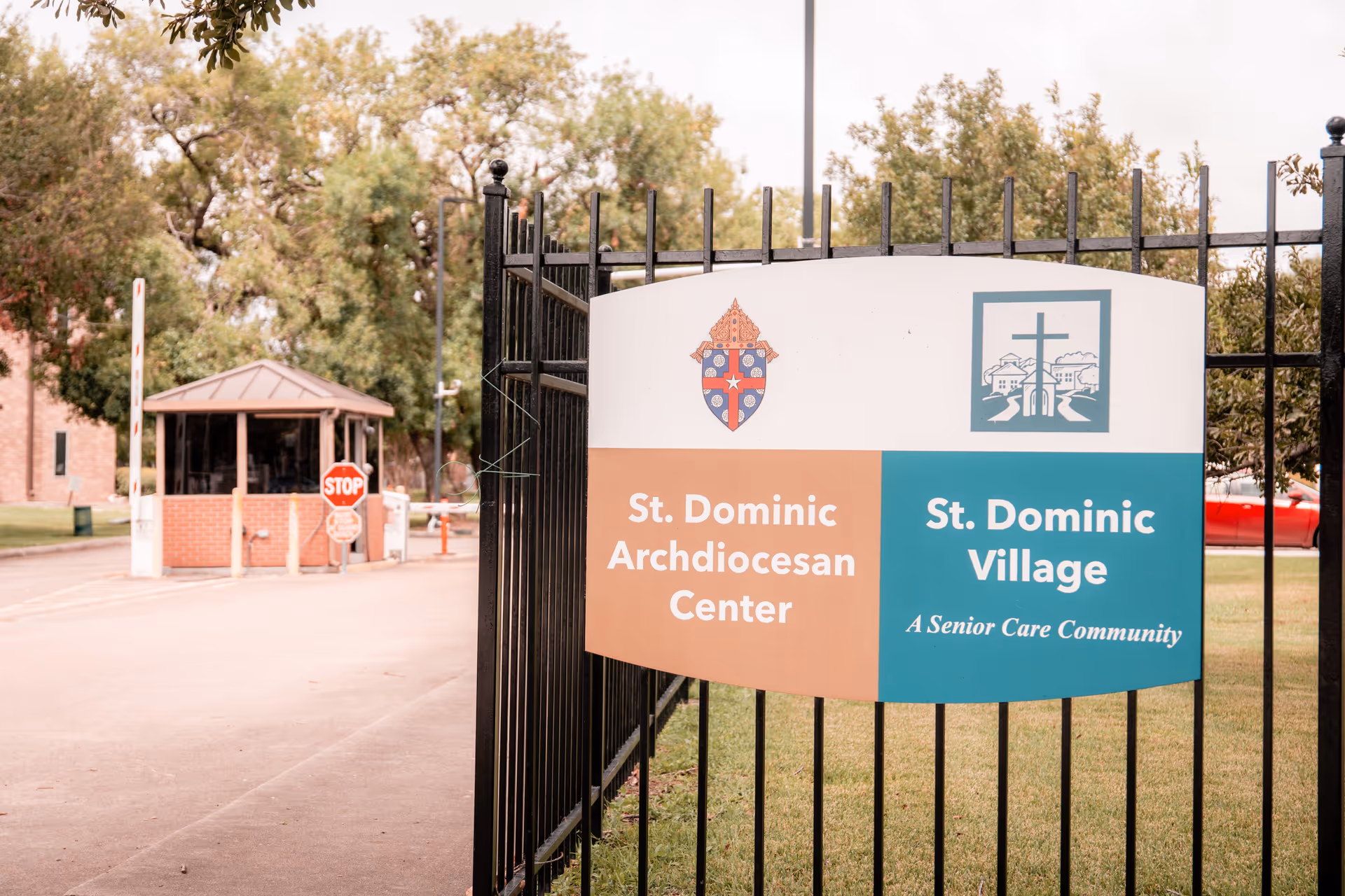 Entrance sign on a black iron fence for St. Dominic Village and St. Dominic Archdiocesan Center with a guardhouse and stop sign visible in the background.