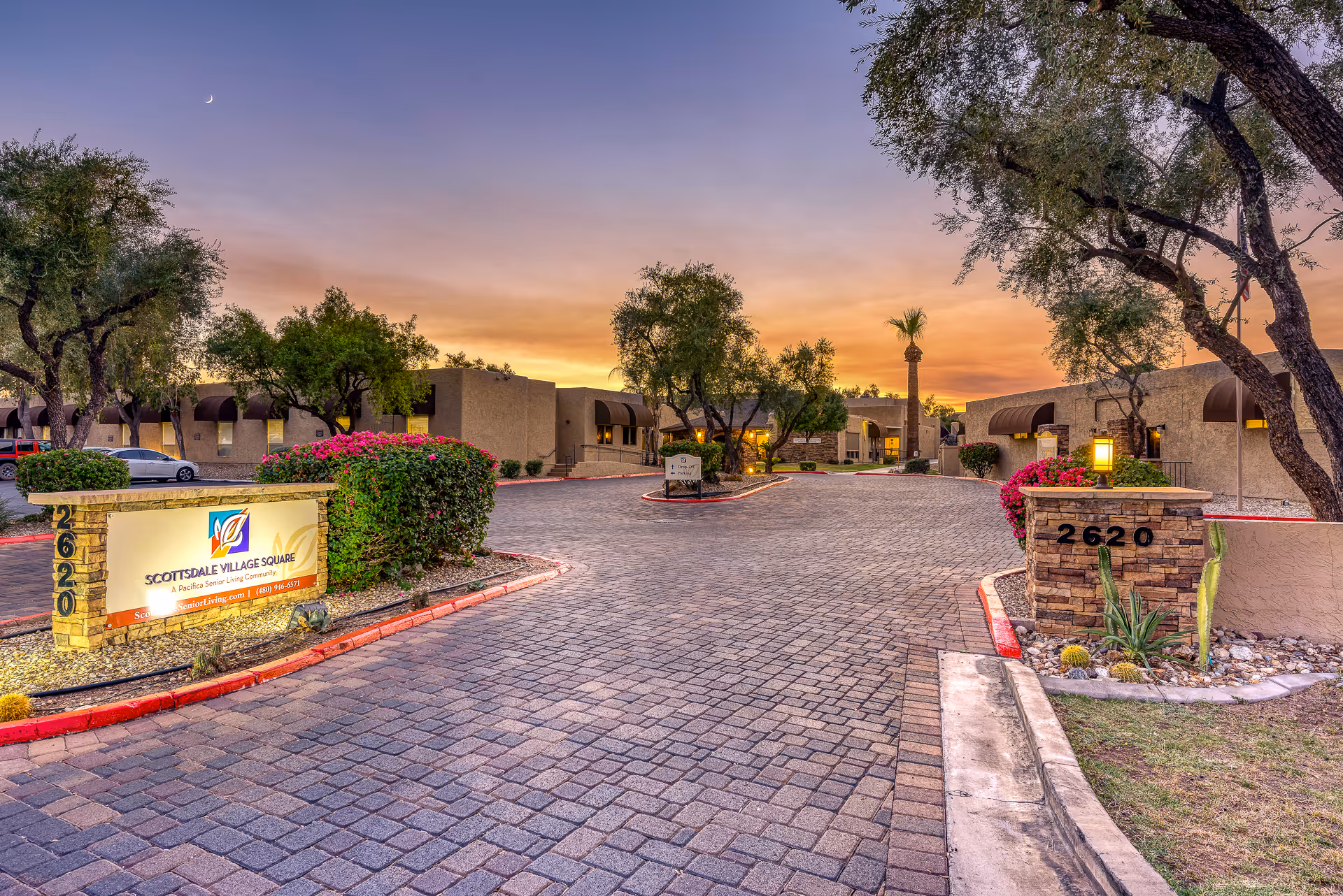 Sunset view of Scottsdale Village Square entrance courtyard with an illuminated sign, low-rise buildings, palm trees, and landscaping.