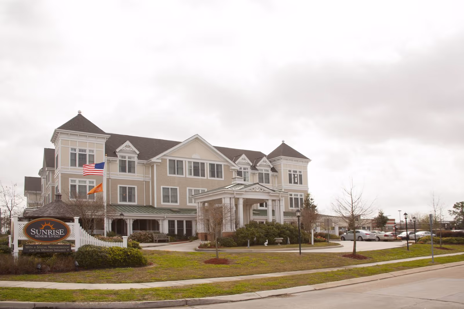 Exterior view of Sunrise Senior Living facility in Metairie, showing a large multi-story building with beige siding, white trim, and a covered entrance. There are two flags on flagpoles near the entrance, a parking lot with cars, and a landscaped lawn with bushes and small trees.
