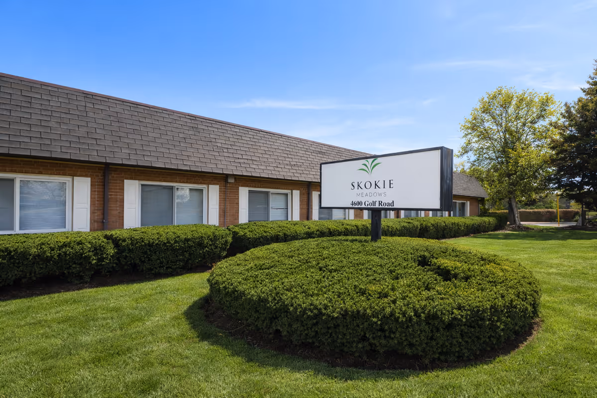 Exterior view of Skokie Meadows facility with sign, shrubs, and blue sky.