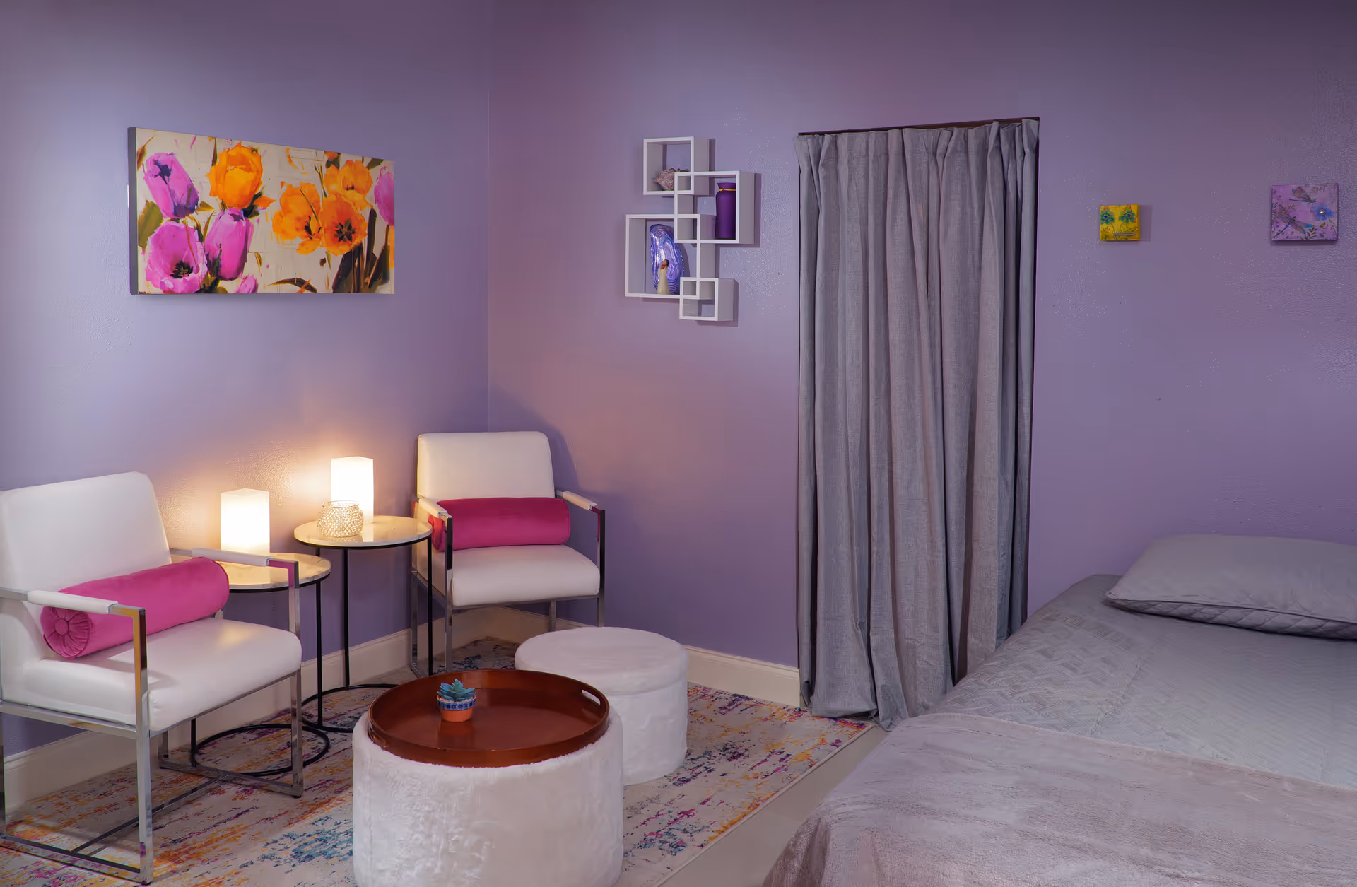 Purple-walled bedroom with two white chairs, lamps, a bed, and decorative art.