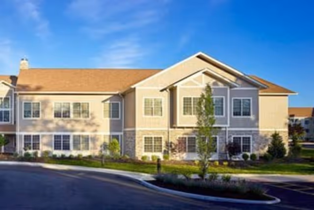 Front exterior of a two-story beige senior living building with multiple windows, landscaped grounds, and a driveway under a blue sky.