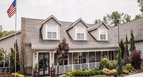 Two-story senior living facility with a wide front porch, landscaping, and an American flag.