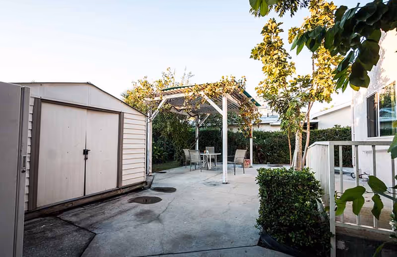 Concrete backyard patio with a storage shed, a pergola-covered seating area, and surrounding greenery.