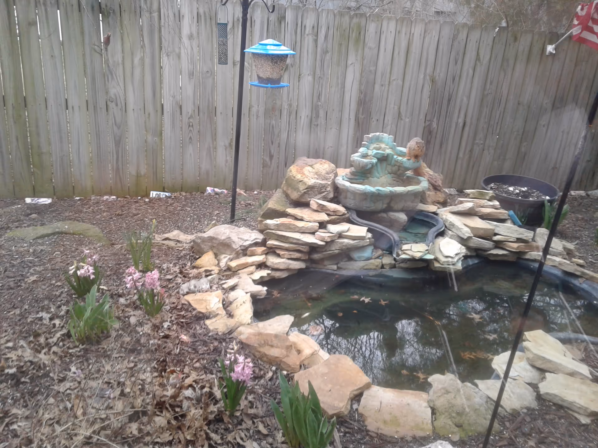 Backyard with a stacked stone pond, small waterfall, bird feeder, and pink flowers along a fence.