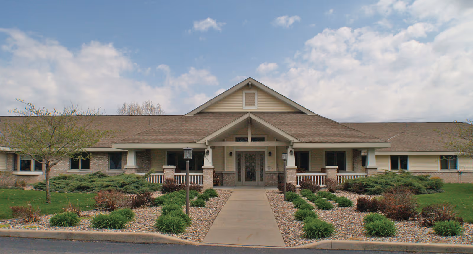 Front exterior of a single-story senior living building with a covered central entrance, walkway and landscaped grounds under a partly cloudy sky.