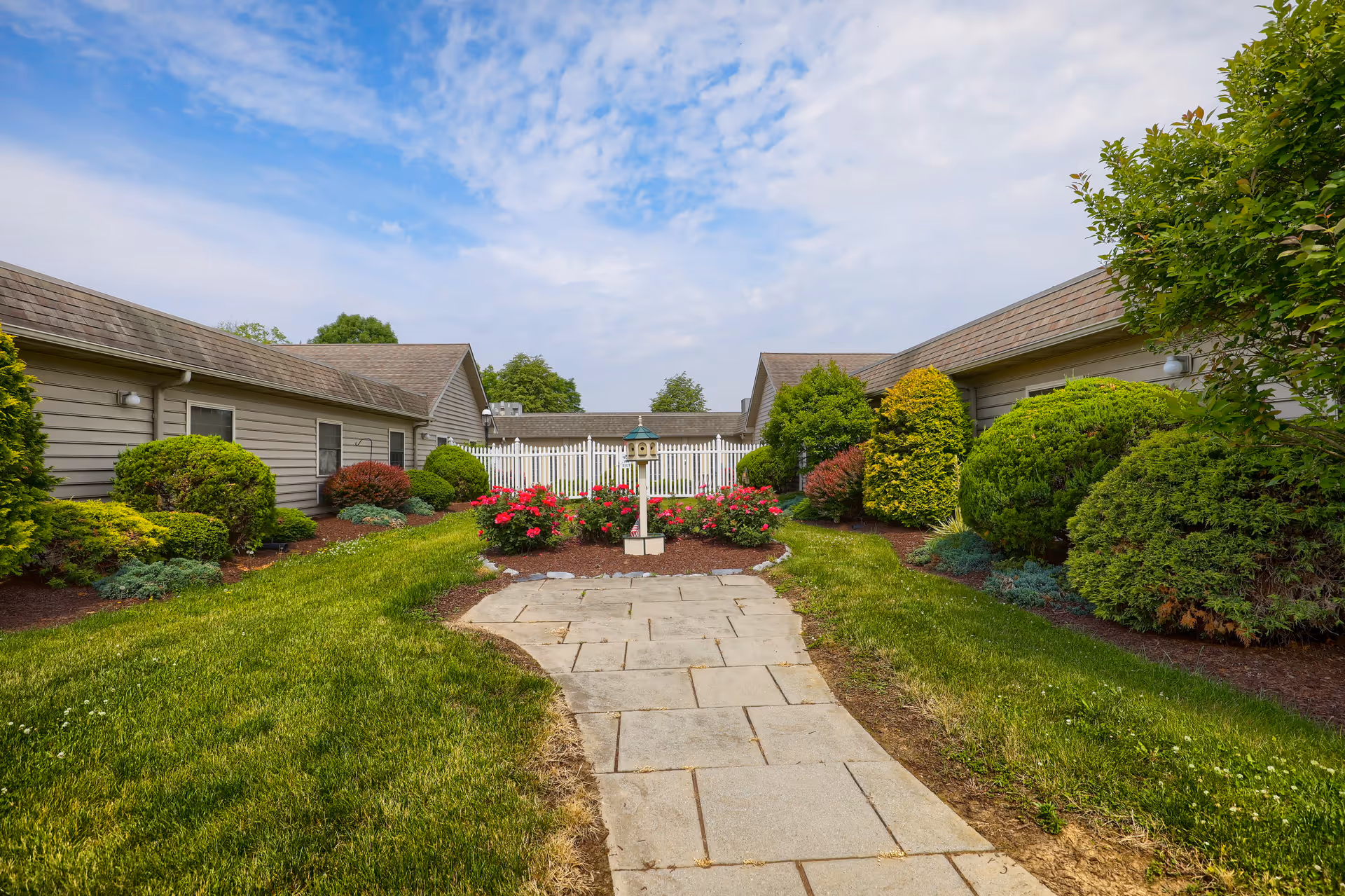 A landscaped outdoor courtyard area with a paved walkway leading to a white picket fence. The courtyard is surrounded by single-story buildings with beige siding and brown roofs. There are well-maintained green bushes, shrubs, and flowering plants along the walkway, under a partly cloudy blue sky.