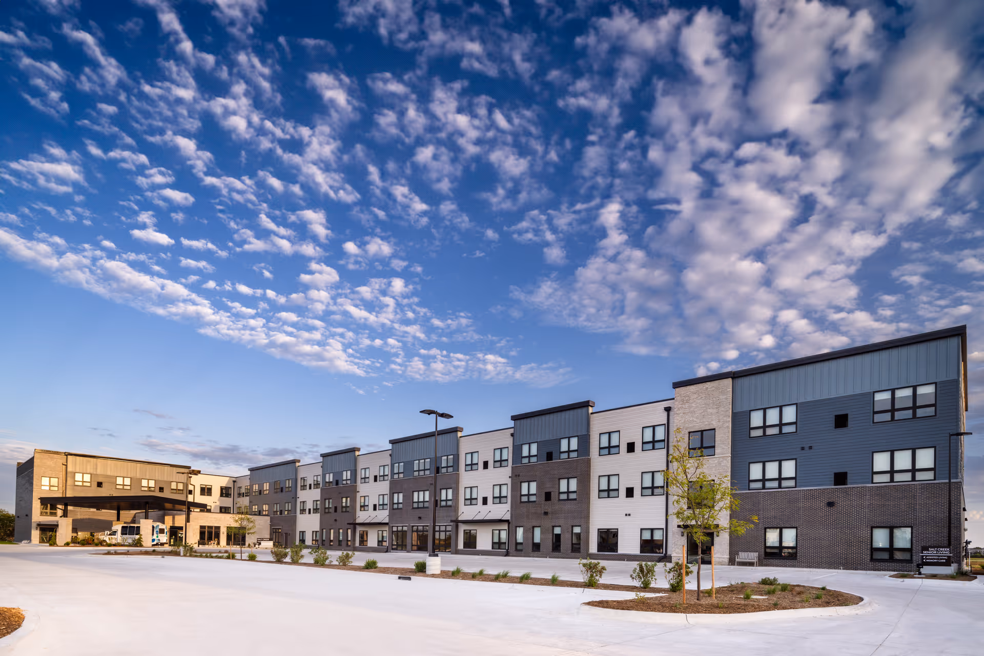 Exterior view of a modern three-story senior living facility building under a partly cloudy blue sky, with a large paved driveway and landscaped areas in front.
