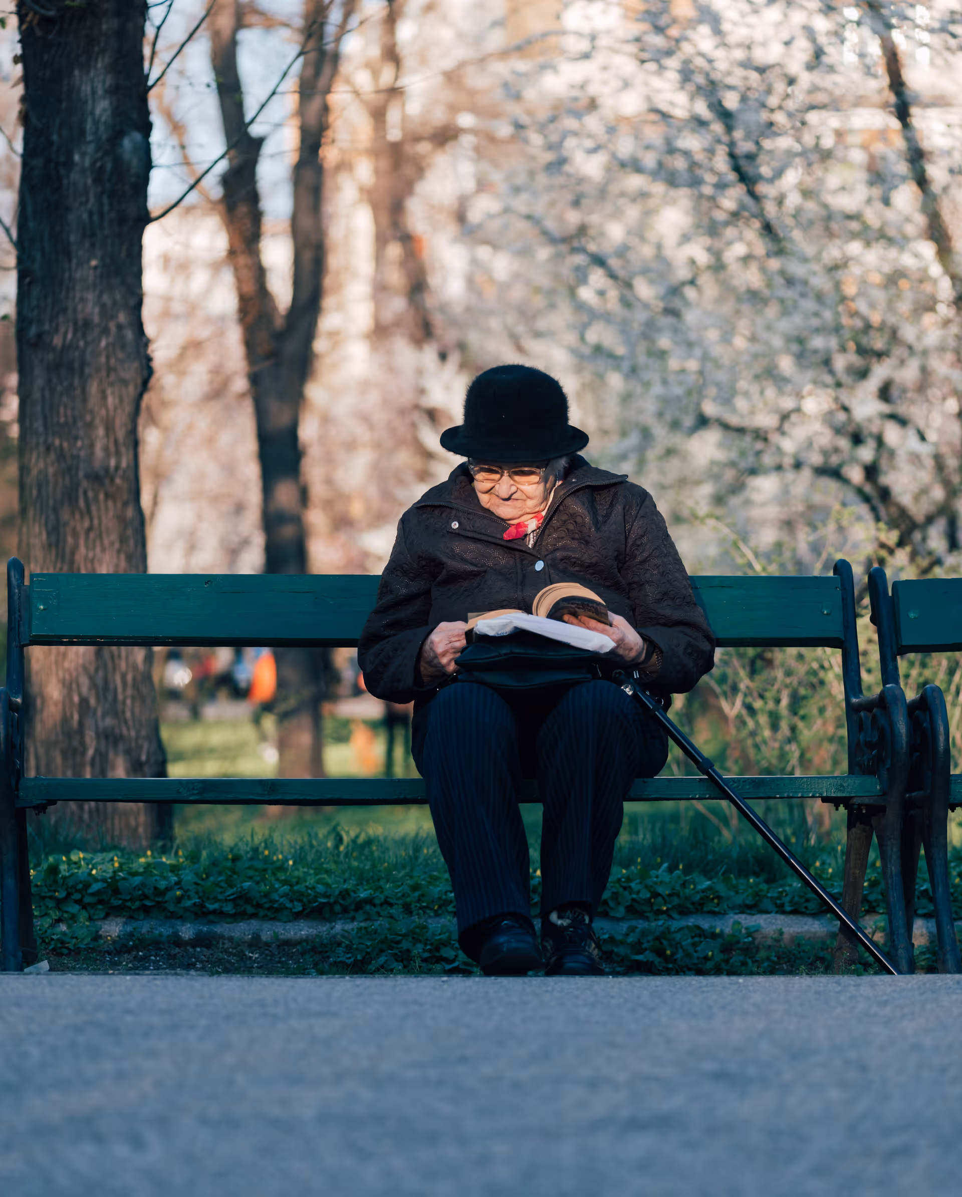 An elderly person sits on a park bench outdoors, reading a newspaper.