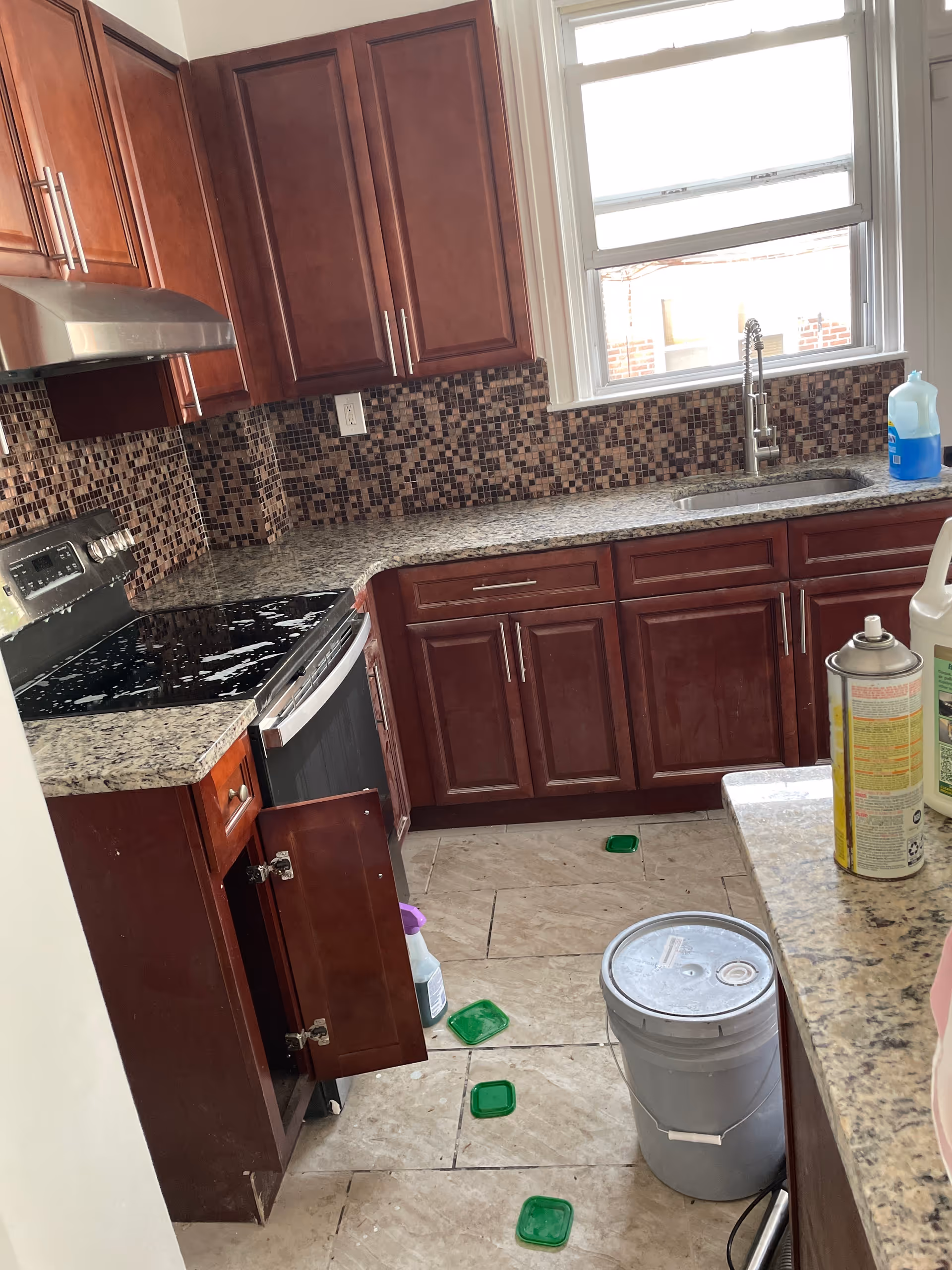 Kitchen with dark wood cabinets, mosaic backsplash, sink, stove, and cleaning supplies.