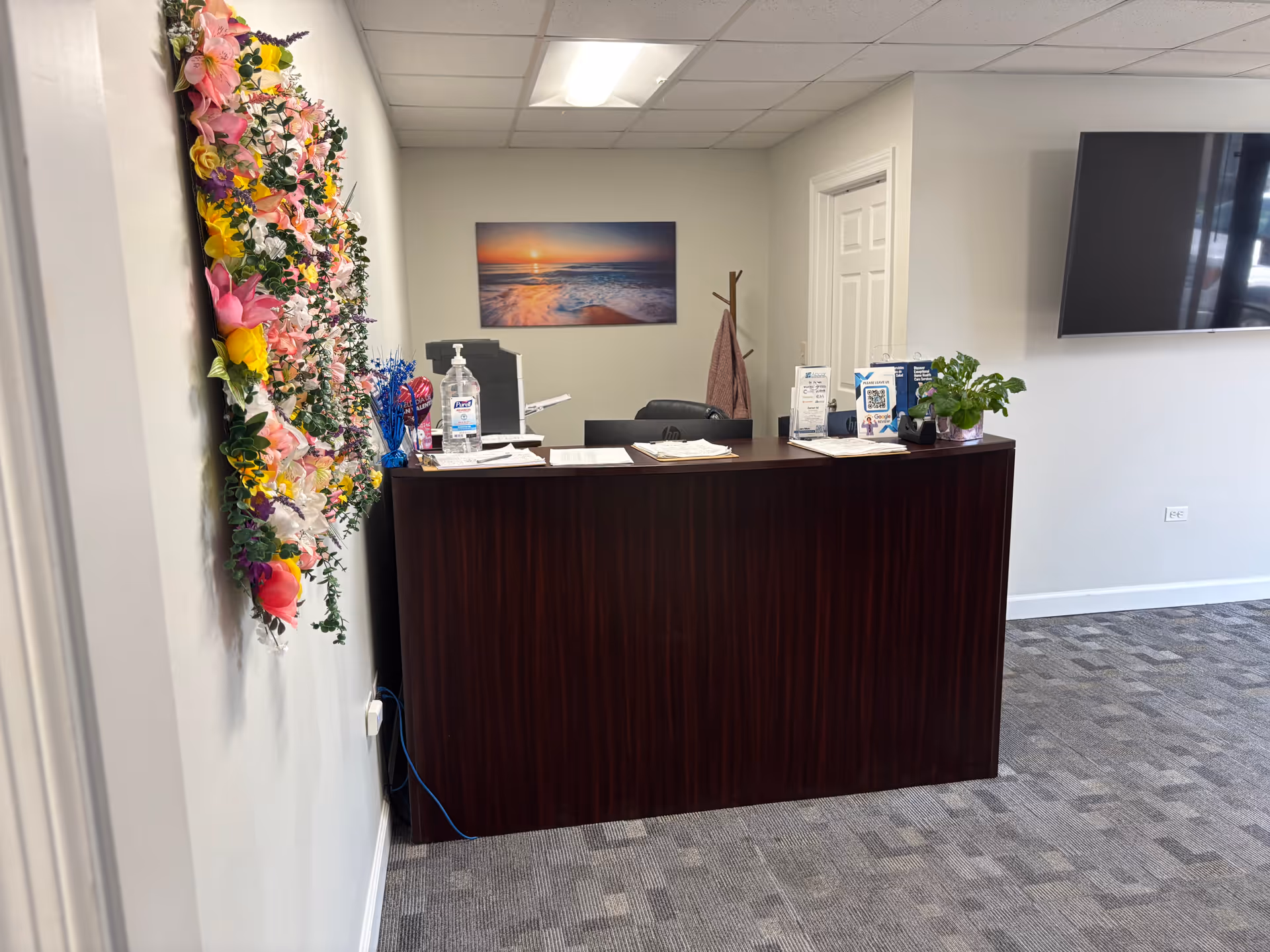 Dark wood reception desk in a bright lobby with wall art and a colorful lei decoration.