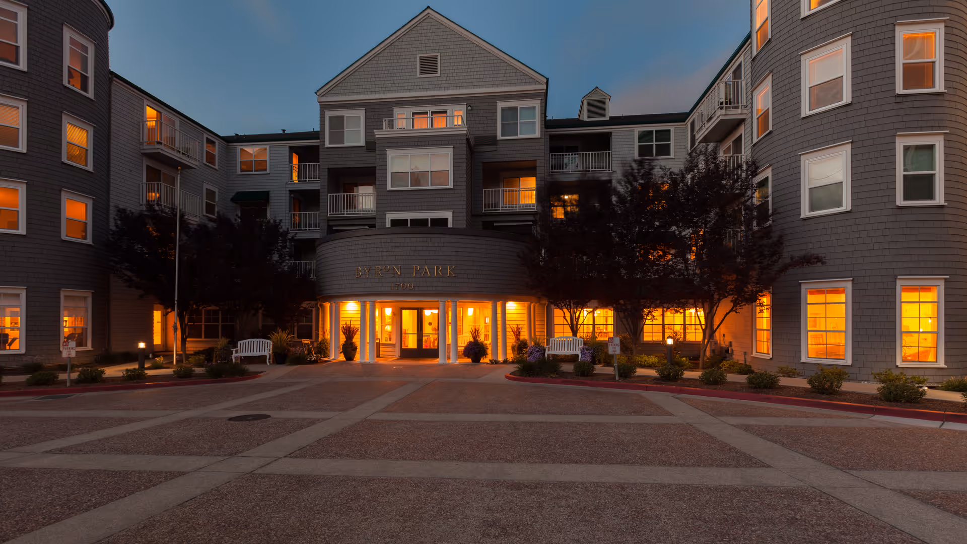 Exterior view of Byron Park senior living facility at dusk with warm lights glowing from the windows. The building has multiple stories with balconies and a central entrance illuminated by outdoor lighting. Trees and benches are visible near the entrance.