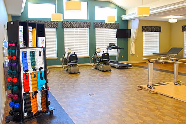 Bright exercise room with recumbent bikes, a treadmill, a rack of weights and therapy tables in a senior living facility.