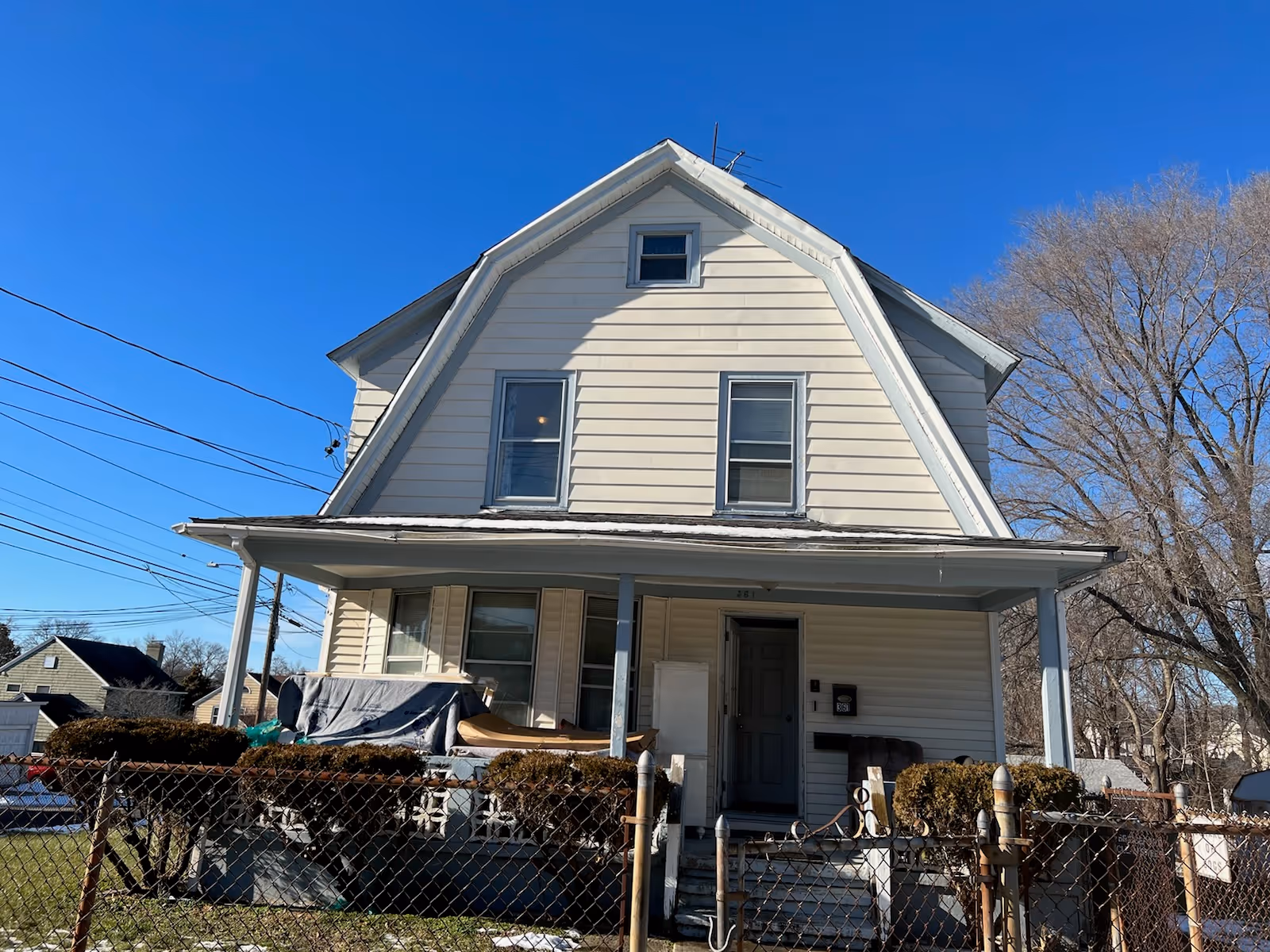 Two-story beige house with a front porch, chain-link fence, and leafless trees.