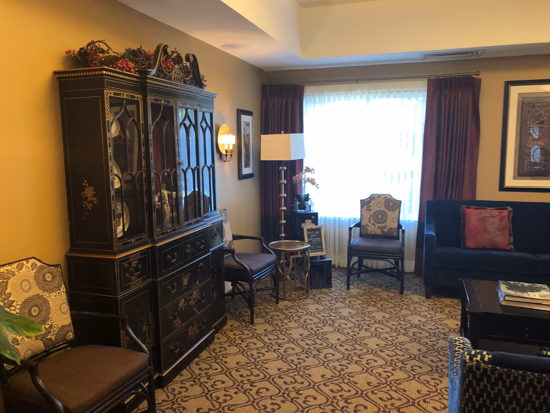 A furnished senior living lounge with dark cabinet, seating, and patterned carpet.