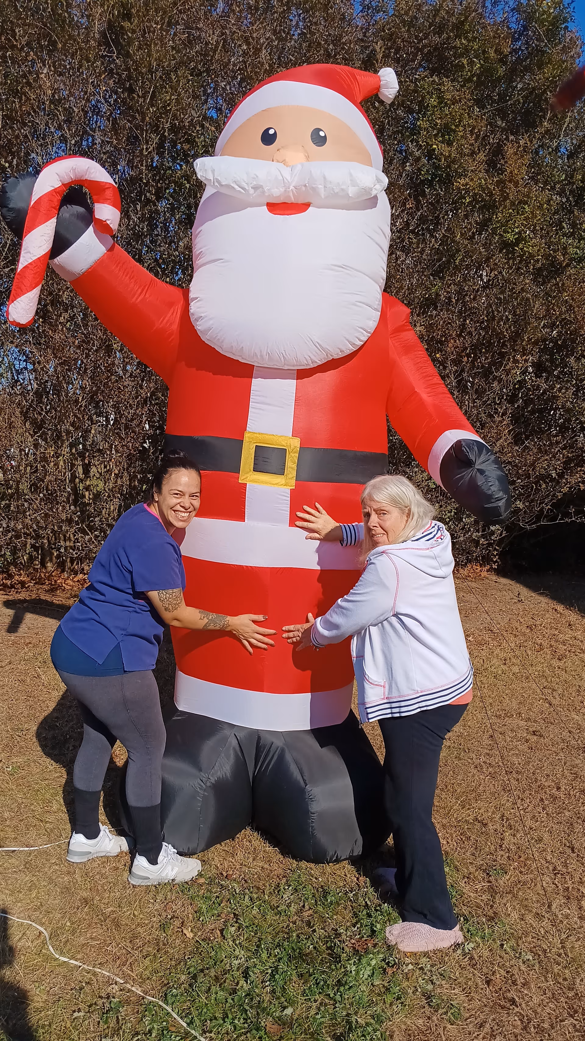 Two women hug a large inflatable Santa Claus outdoors beside a grassy area.
