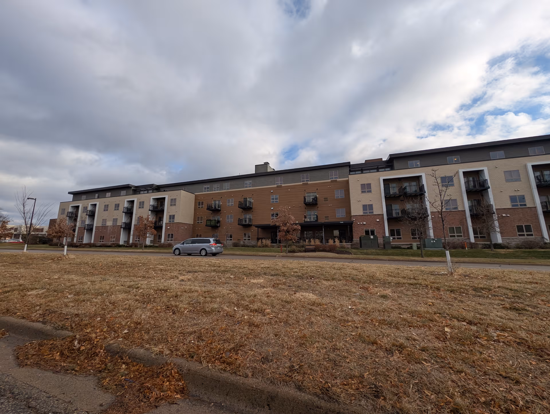 Exterior view of a multi-story senior living facility building with balconies, a parked silver minivan, leafless trees, and a cloudy sky.
