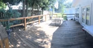 A spacious wooden outdoor deck area with a wooden railing on the left side and several tables and chairs arranged on the deck. The deck is adjacent to a white building with large windows and doors. Trees and greenery are visible beyond the railing, providing a shaded and serene environment.