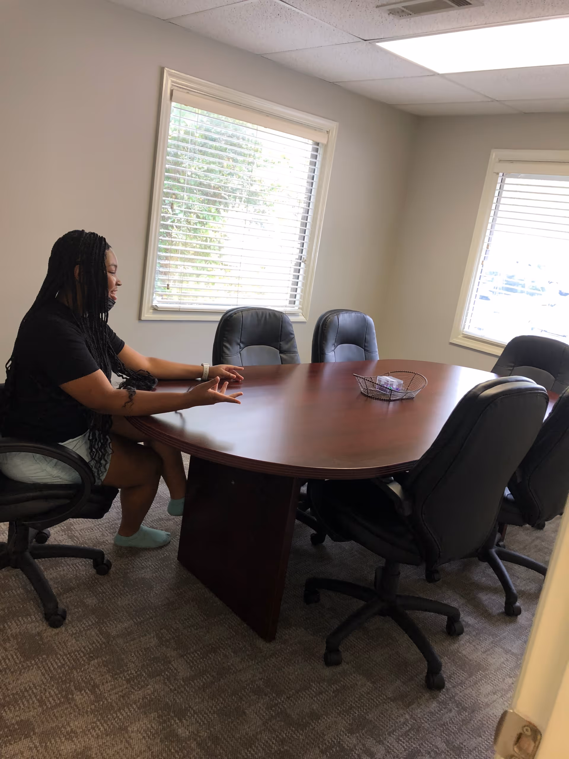A woman sits at a round wooden conference table in a bright, windowed meeting room.