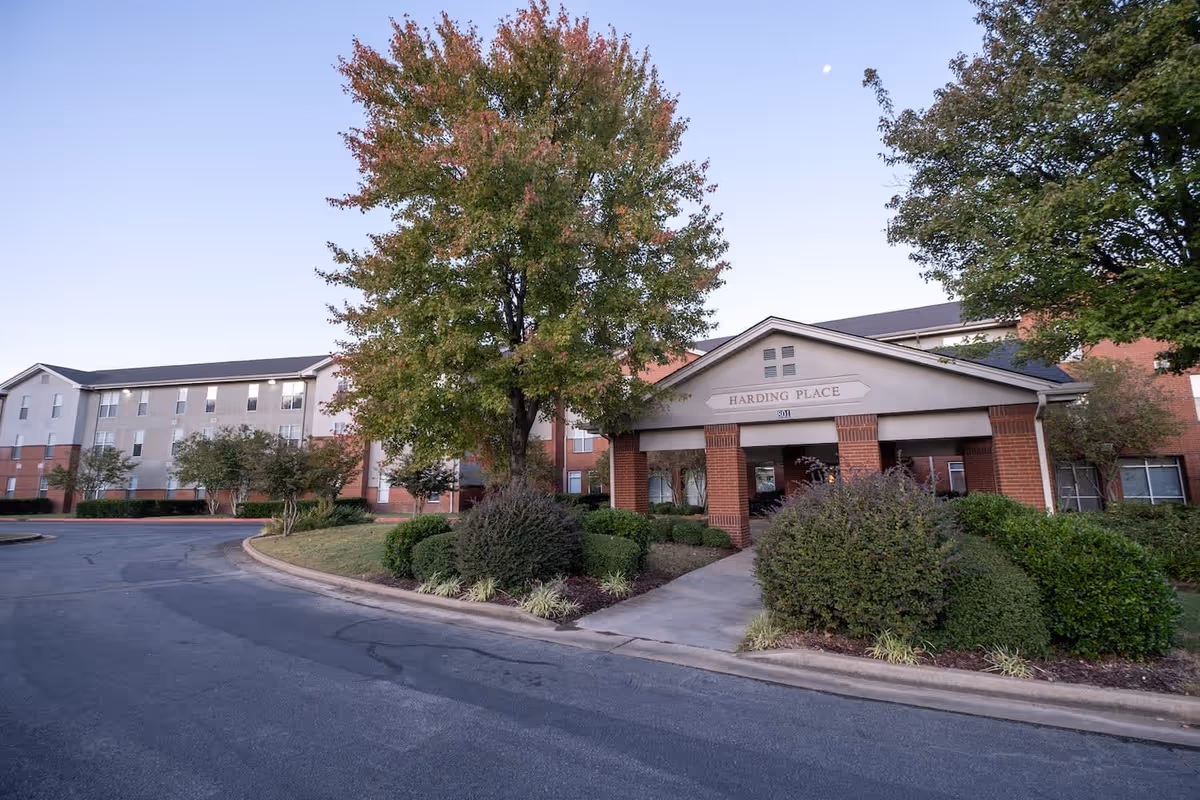 Exterior view of Harding Place Retirement facility showing a multi-story building with a covered entrance, surrounded by trees and landscaped bushes under a clear sky.