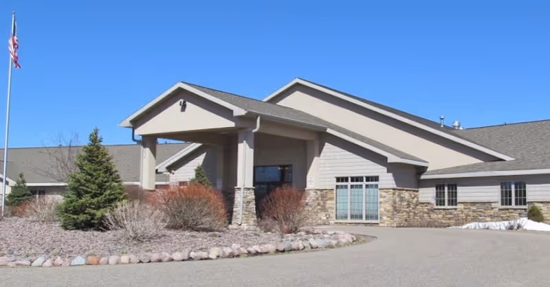 Front entrance of a single-story senior living building with a covered porte-cochere, stone facade, and landscaped circular driveway under a clear blue sky.
