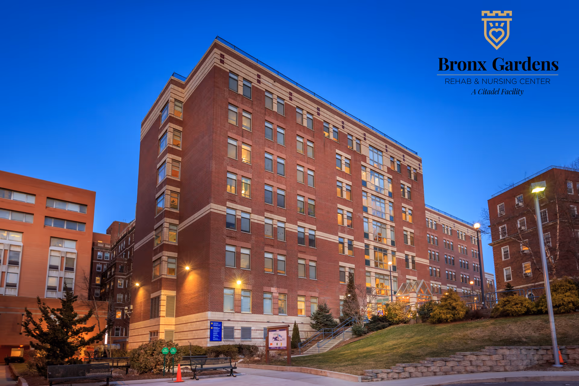 Large brick senior living facility exterior with Bronx Gardens logo against dusk sky.