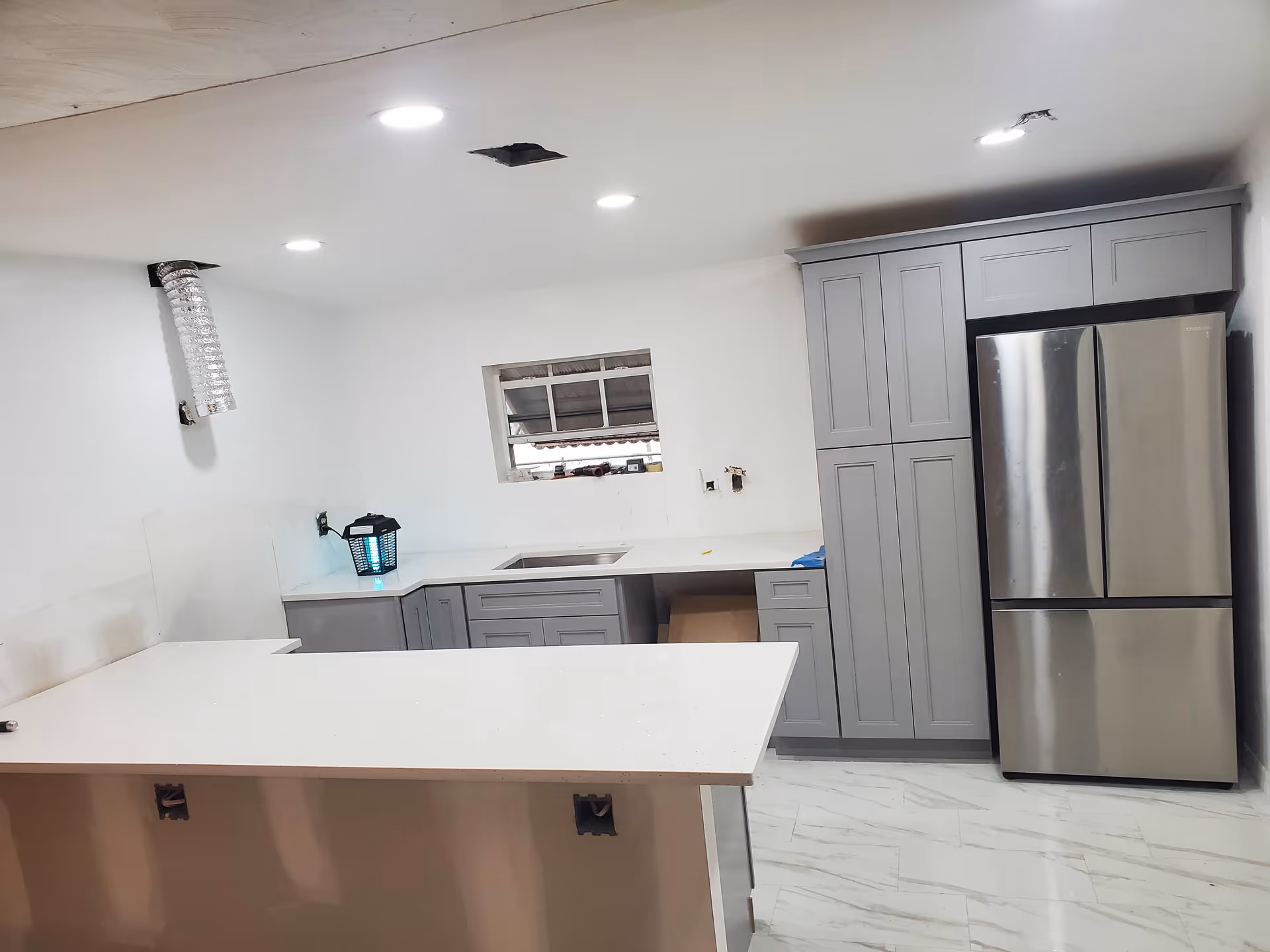 Modern kitchen with gray cabinets, white countertops, stainless fridge, and ceiling lights.