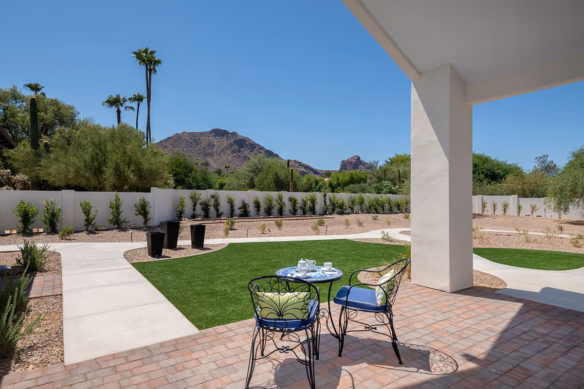 Covered patio with a small table and two chairs overlooking a landscaped yard with artificial turf, desert plants, and mountains in the distance.