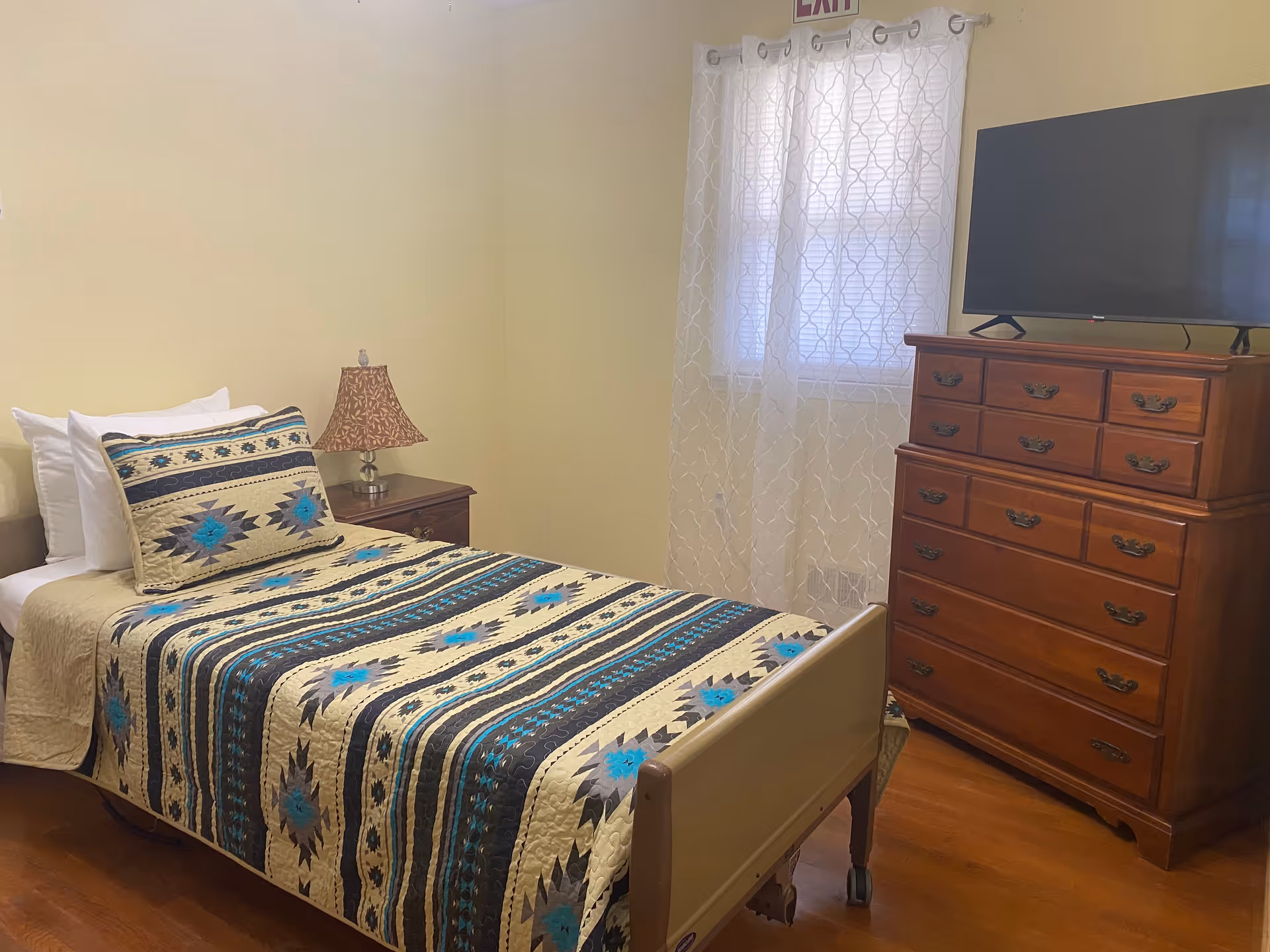 Cozy bedroom with bed, dresser, lamp, TV, and curtained window.