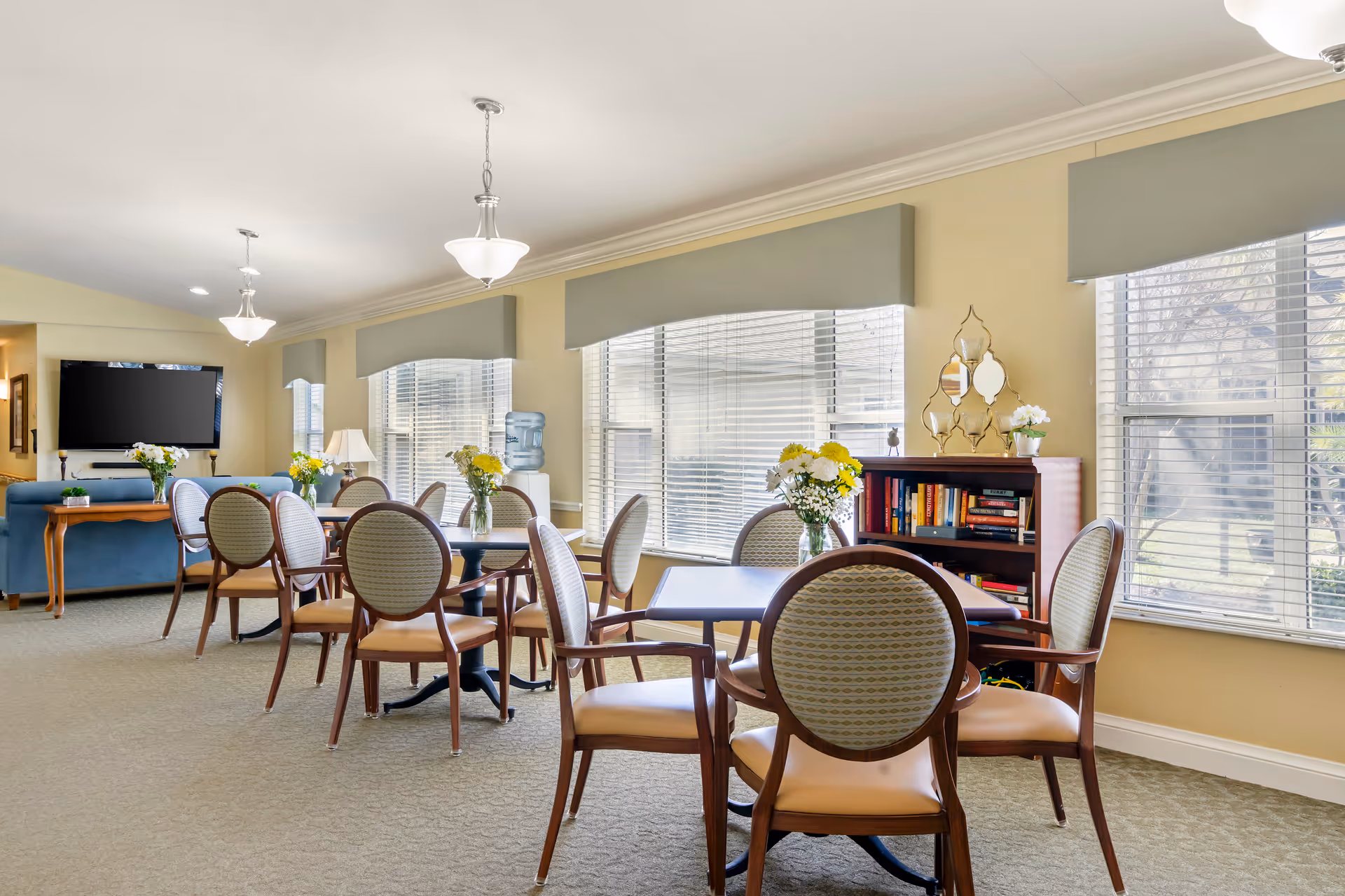 Bright senior living dining room with round tables, chairs, windows, and flowers.