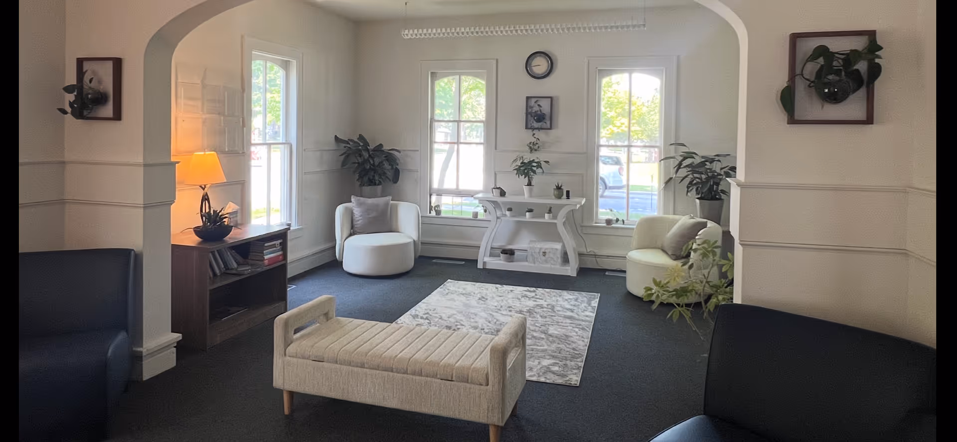 Sunlit living room with white seating, plants, a central console, and windows.