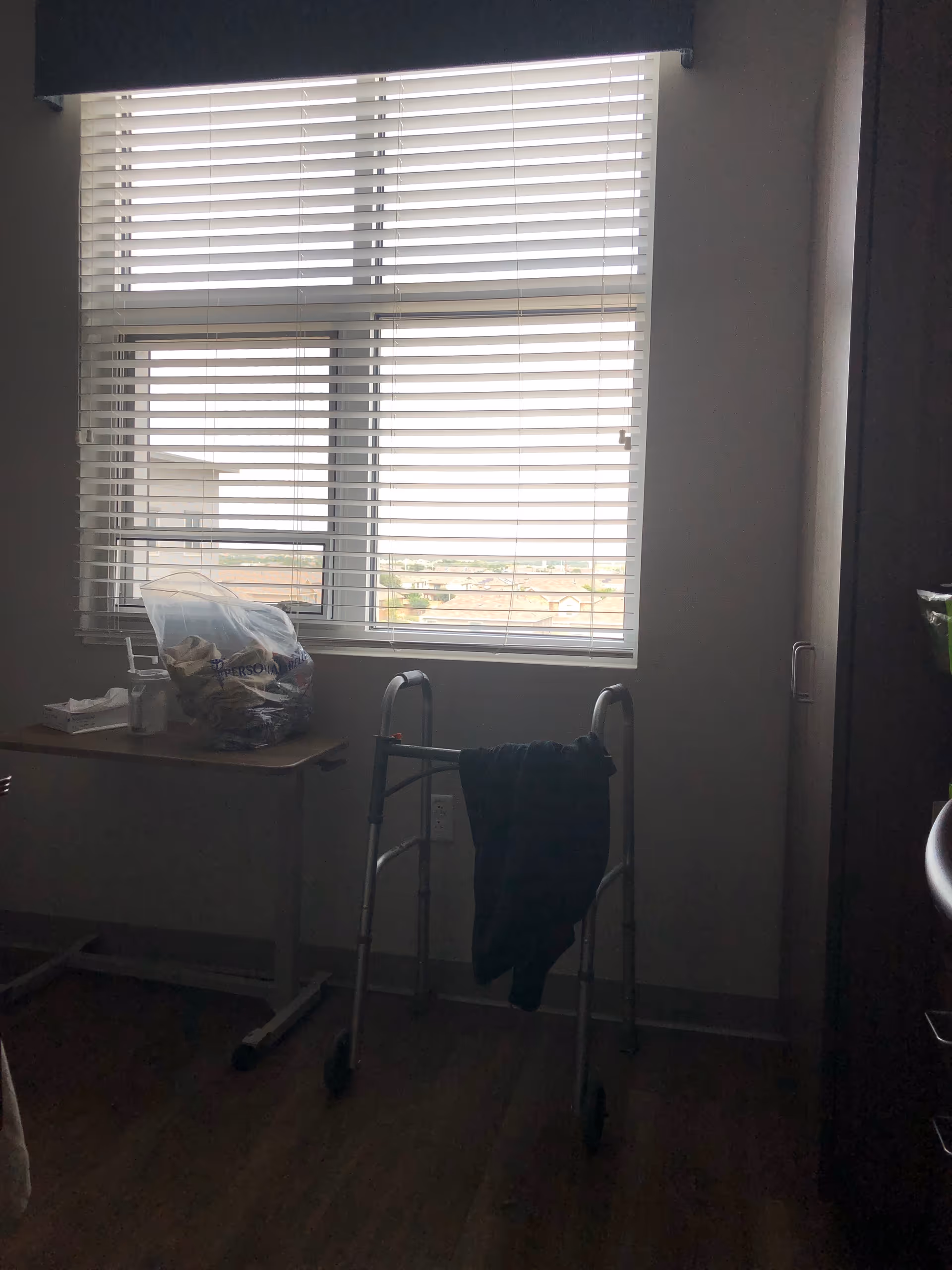 A walker stands by a desk near a window with closed blinds in a dim room.