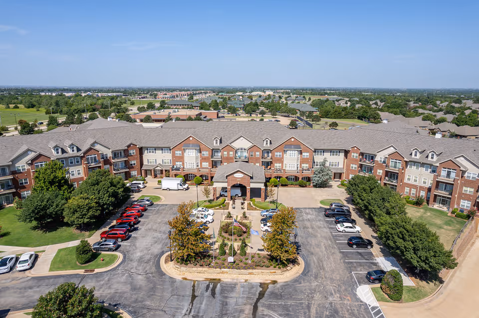 Aerial view of a large senior living facility building with a U-shaped layout, surrounded by parking lots with cars, landscaped greenery, and trees under a clear blue sky.