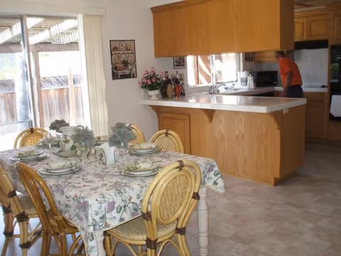 A dining room scene with a floral tablecloth set for a meal and a kitchen beyond.