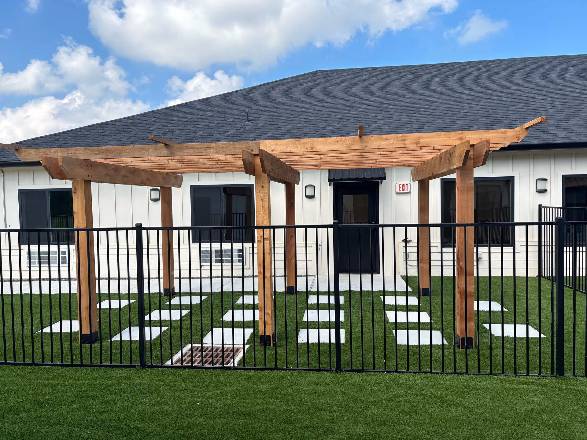 Outdoor area of a senior living facility with a wooden pergola structure over a green artificial lawn. There are white stepping stones arranged in a pattern on the grass, a black metal fence in the foreground, and a building with white siding and black-framed windows and doors in the background under a partly cloudy sky.