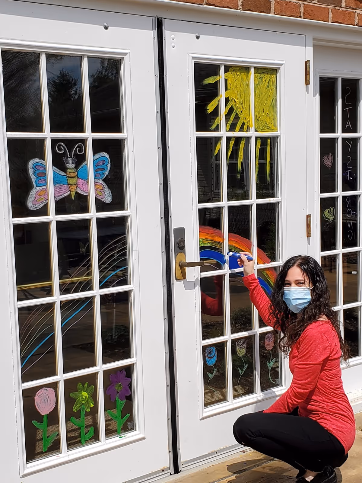 Person wearing a mask paints a rainbow on glass doors at a senior living facility entrance.