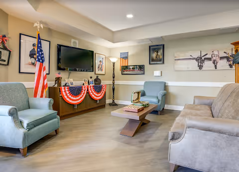 Cozy senior living lounge with blue chairs, sofas, a TV, and patriotic decor.
