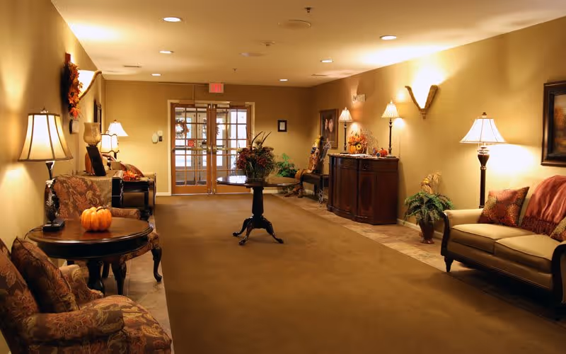 A warmly lit hallway in a senior living facility with comfortable seating areas on both sides, decorated with table lamps, plants, framed artwork, and autumn-themed decorations including a small pumpkin on a side table. Double glass doors are visible at the end of the hallway.