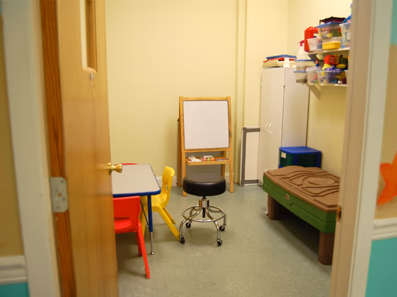 Child-sized furniture, play table and chairs, a whiteboard, and storage shelves in a therapy room.
