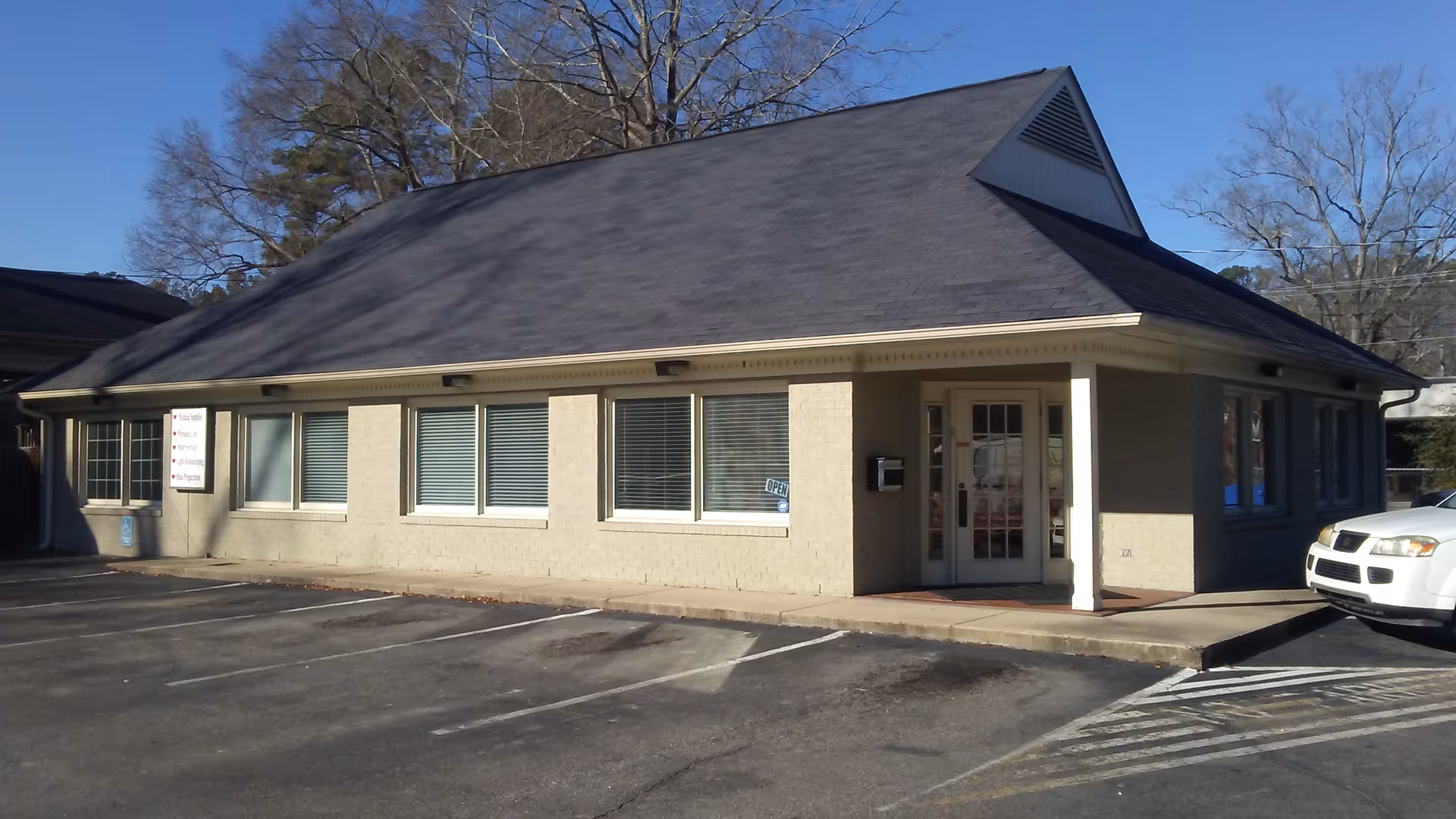 Single-story beige building with a dark roof, large windows, and a parking lot in front.