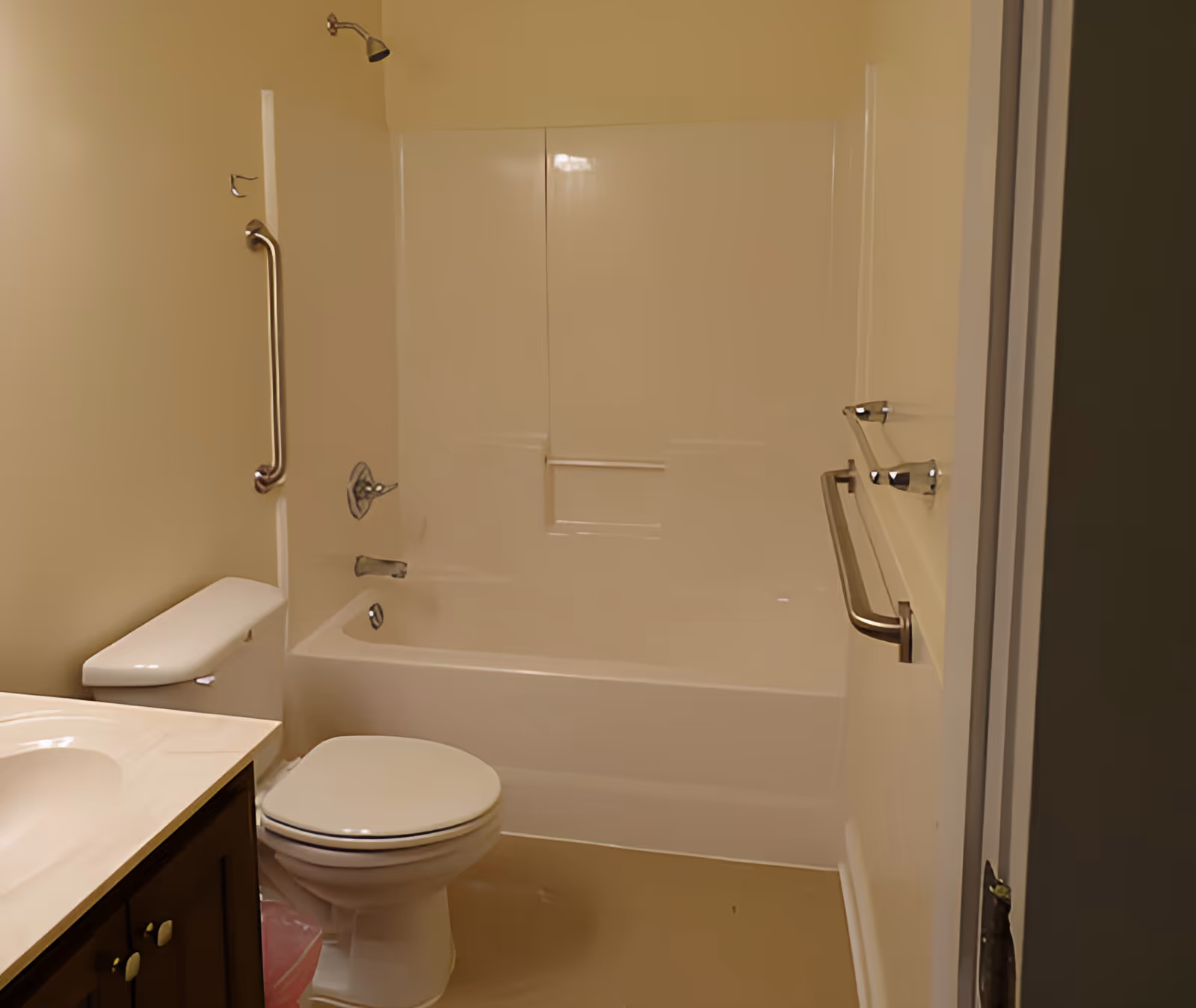 A bathroom with a white bathtub and shower combination, a toilet, a sink with a countertop, and grab bars on the wall near the bathtub and beside the toilet. The walls are painted a light beige color.