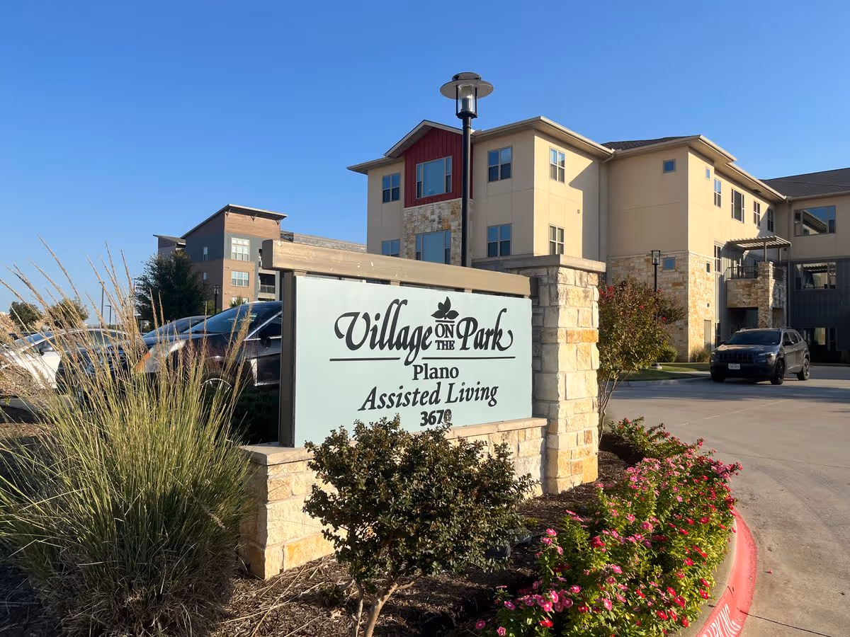 Entrance sign reading 'Village on the Park Plano Assisted Living' in front of the facility building and landscaped driveway.
