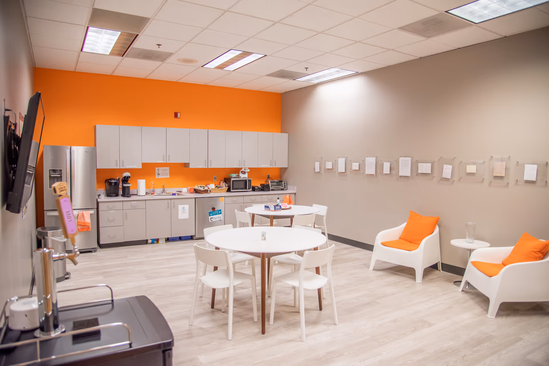 Bright kitchenette and lounge area with orange accent wall, white cabinets, tables, and chairs.