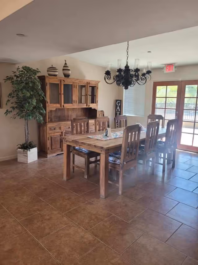 Wooden dining table with six chairs, a matching hutch, chandelier, potted plant, and glass doors in a bright dining room.
