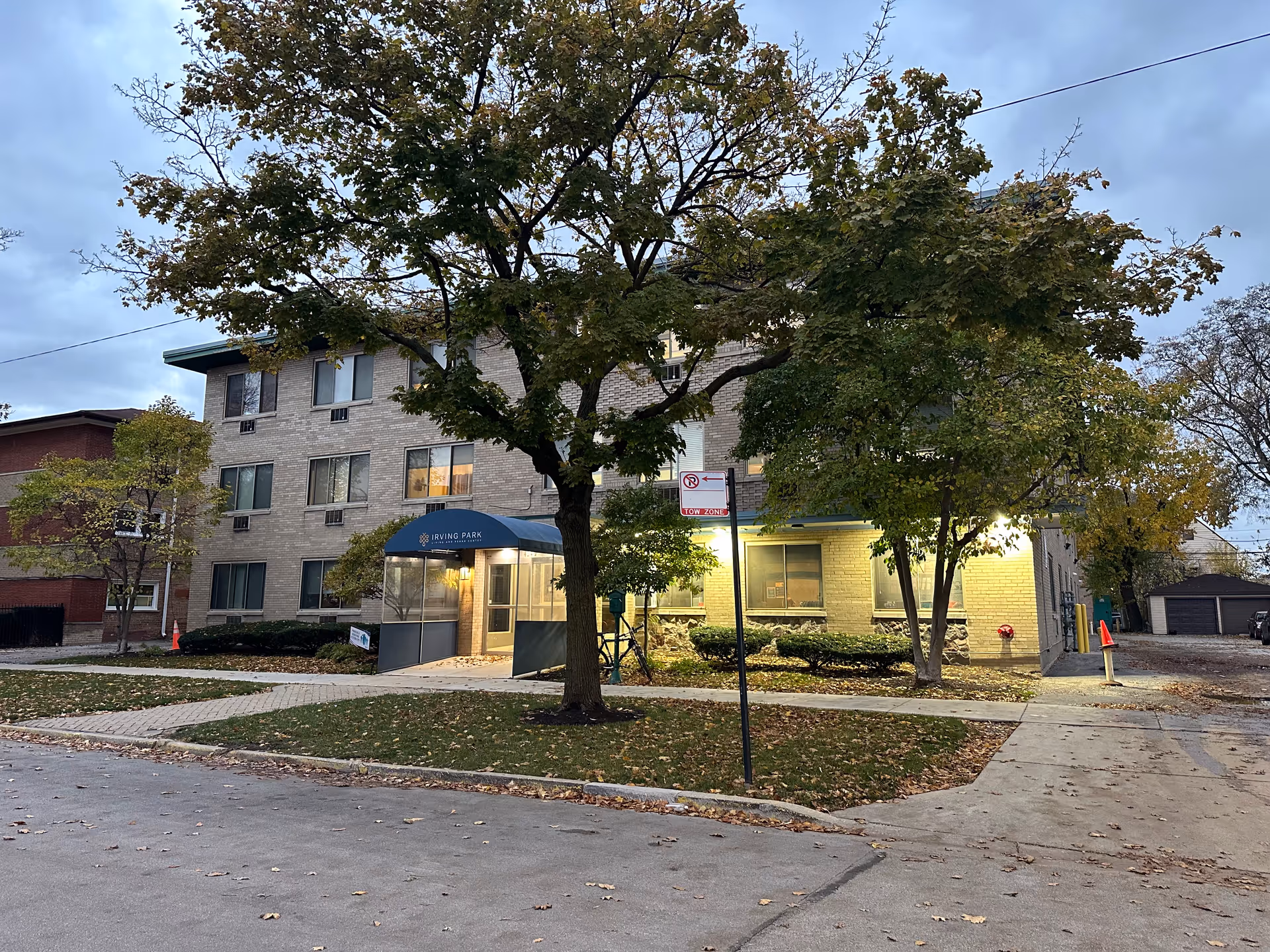 Exterior view of a three-story brick building with a blue awning over the entrance that reads 'Irving Park'. The building is surrounded by trees with green and yellow leaves, and there is a sidewalk and street in the foreground. It appears to be early evening with lights on near the entrance.