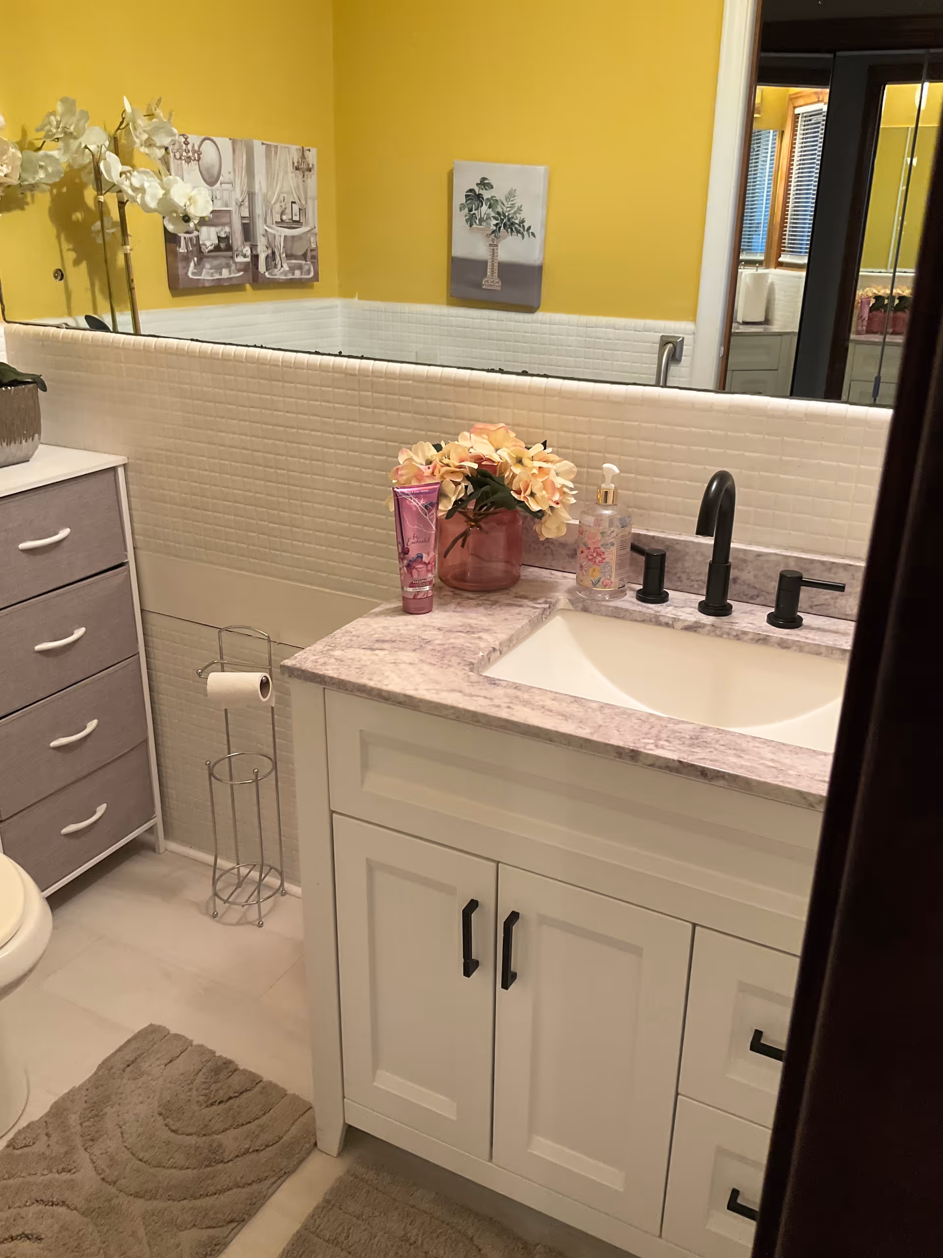 Bright bathroom with white vanity, marble countertop, yellow walls, and decorative flowers.