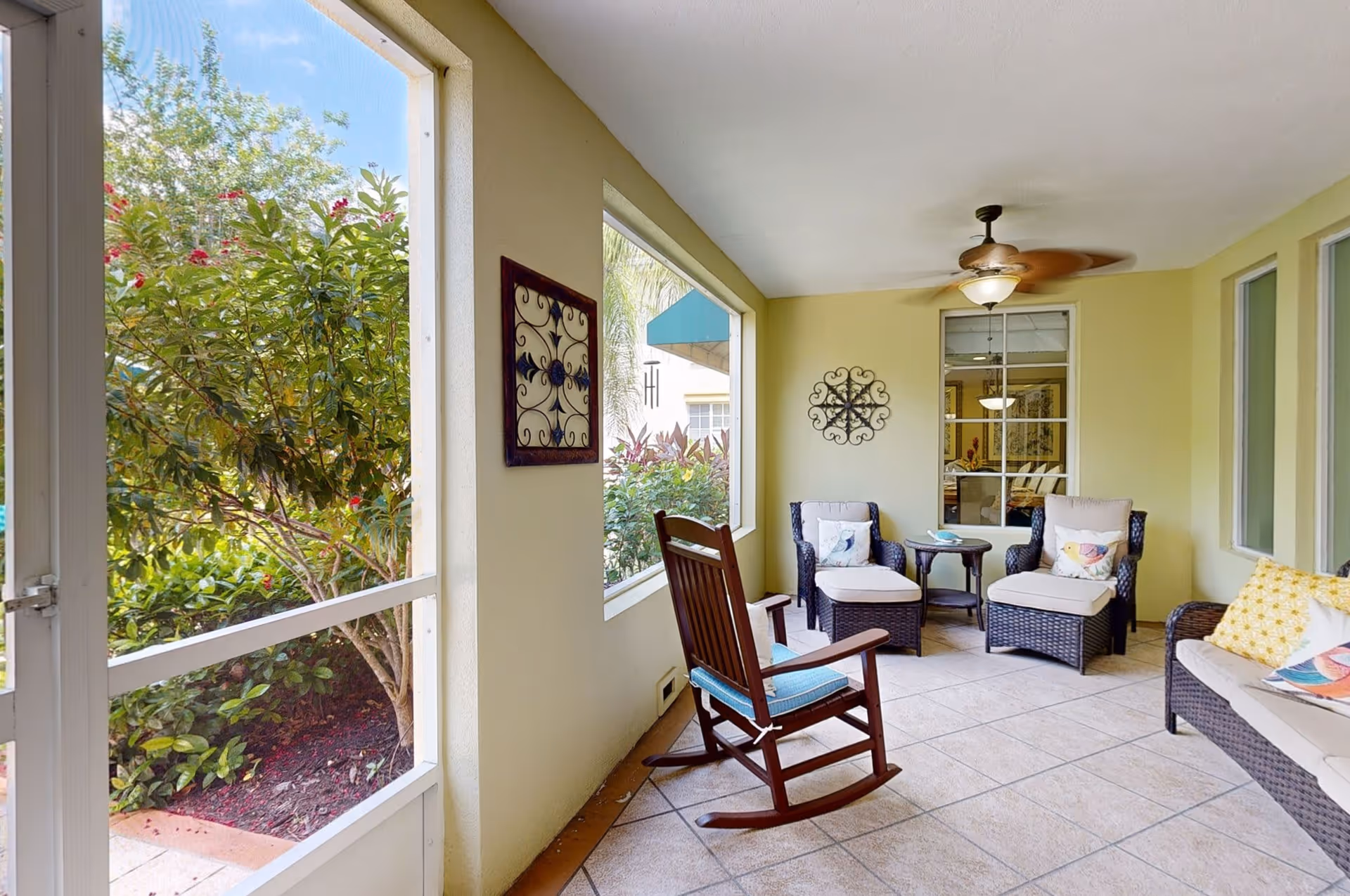 A cozy screened-in porch area with a ceiling fan, featuring a wooden rocking chair with a blue cushion, two wicker armchairs with cushions and pillows, a small round table, and a wicker sofa with colorful pillows. Outside the porch, there are green bushes and trees with red flowers.