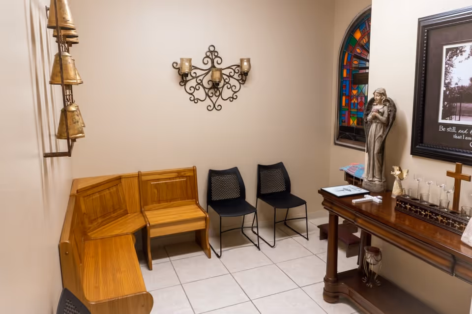 Small, quiet chapel-like room with wooden benches, two chairs, altar, statue, and stained-glass window.
