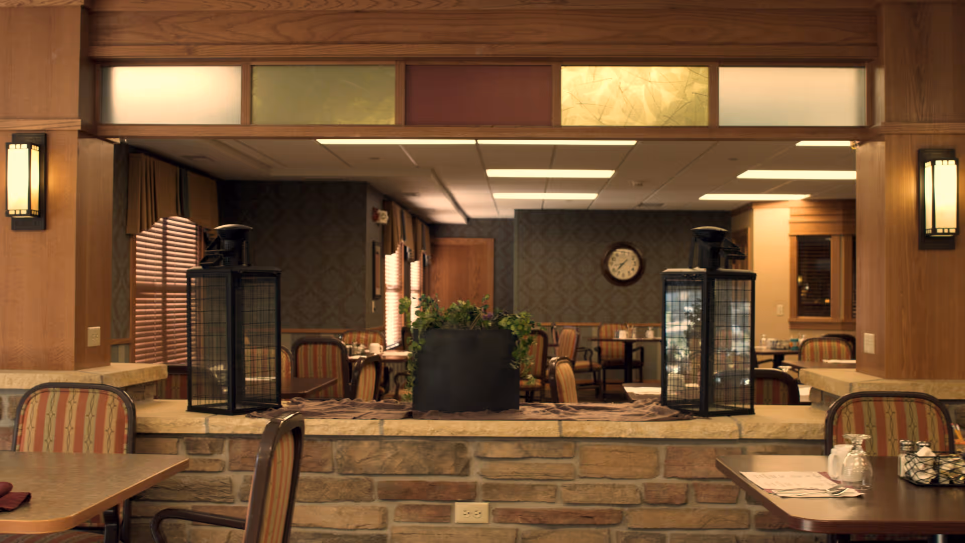 Interior view of a dining area in a senior living facility with wooden paneling, stone half-wall, and tables with striped cushioned chairs. Two large black lanterns and a potted plant are placed on the stone half-wall. The room has patterned wallpaper, a clock on the wall, and windows with wooden blinds.